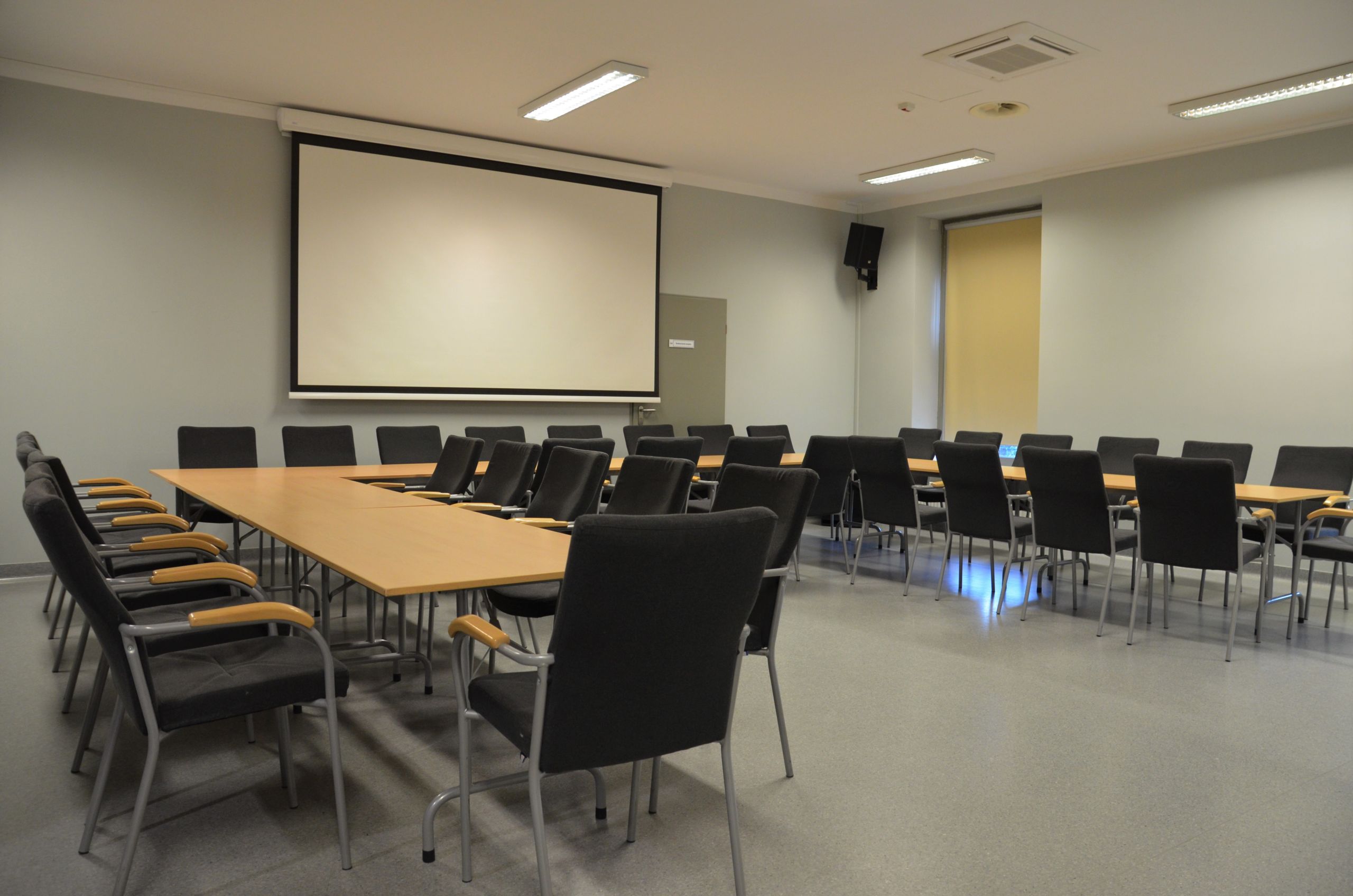 Zdjęcie przedstawiające salę konferencyjną. Przedstawia stoły ułożone w kształt litery C. Po obu stronach stołu poustawiane są krzesła. Na ścianie znajduje się ekran do rzutnika, a w rogach sali znajdują się głośniki.