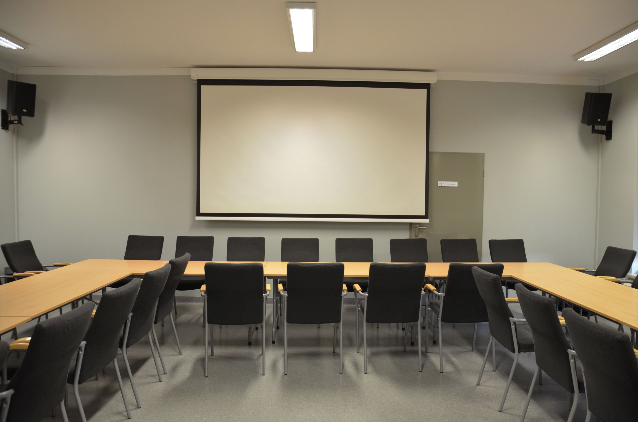 Zdjęcie przedstawiające salę konferencyjną. Przedstawia stoły ułożone w kształt litery C. Po obu stronach stołu poustawiane są krzesła. Na ścianie znajduje się ekran do rzutnika, a w rogach sali znajdują się głośniki.