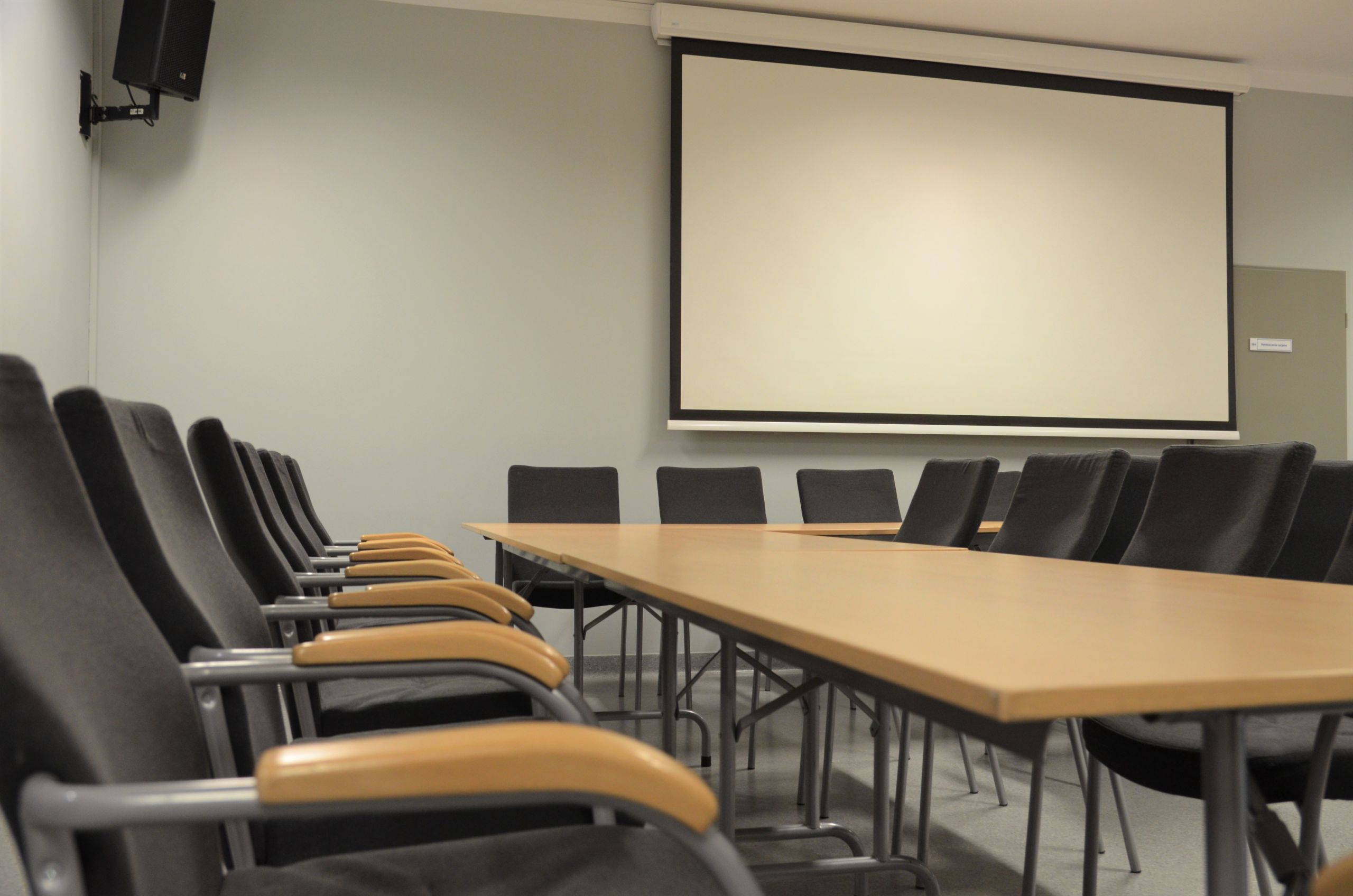 Zdjęcie przedstawiające salę konferencyjną. Przedstawia stoły ułożone w kształt litery C. Po obu stronach stołu poustawiane są krzesła. Na ścianie znajduje się ekran do rzutnika, a w rogu sali znajduje się głośnik.