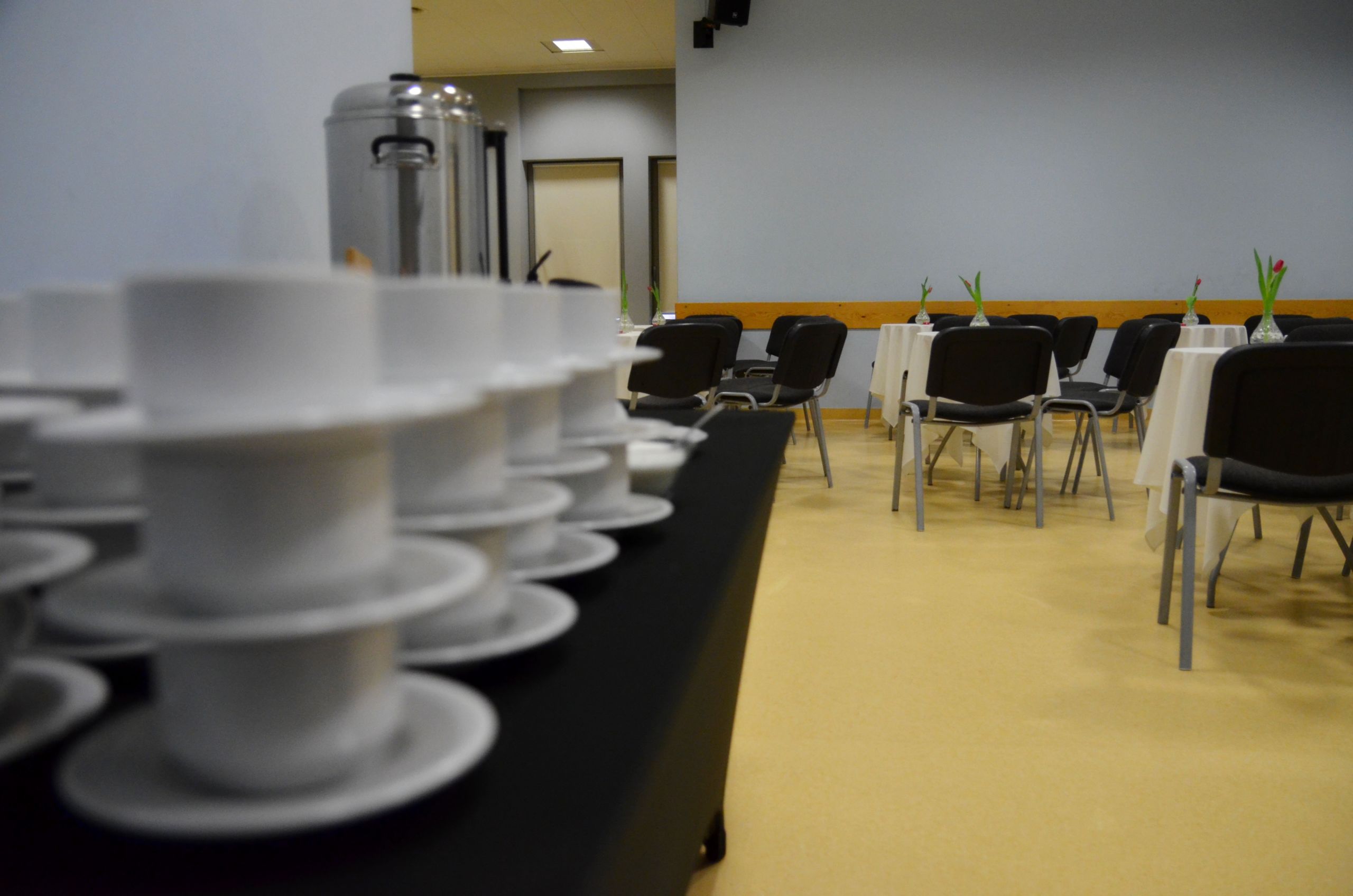 Zdjęcie przedstawiające salę kameralną - po lewej stronie znajduje się blat okryty czarnym obrusem. Na nim znajdują się poustawiane białe filiżanki oraz ekspresy do kawy oraz pojemniki z wodą. Po przeciwnej stronie sali znajdują się okrągłe stoły z krzesłami dookoła.