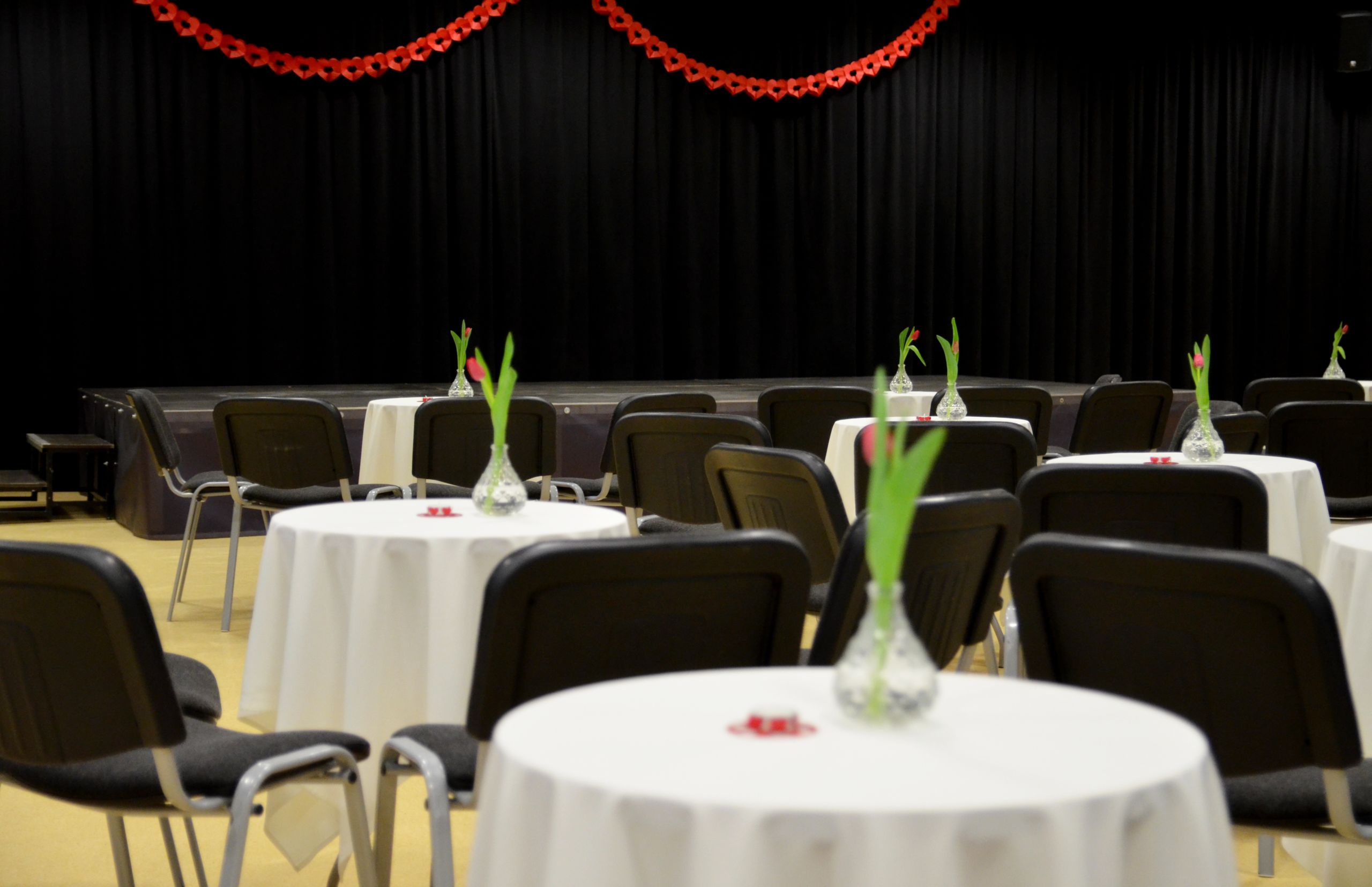 Zdjęcie przedstawiające salę kameralną. Wypełniona jest okrągłymi stołami i białymi obrusami oraz czarnymi krzesłami dookoła. Na każdym stole postawiony jest wazon z tulipanem. W tle znajduje się podest - scena z czarną kurtyną.