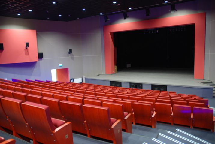 Zdjęcie przedstawiające wnętrze sali kinowo-widowiskowej MOK Międzyrzecz. Sala wypełniona jest rzędami czerwonych foteli kinowych. Po obu stronach sali prowadzą do nich rzędy podświetlonych ledami schodów. Przed fotelami znajduje się podwyższona scena. Na niej znajduje się wejście na kulisy w miejscu, w którym w trakcie projekcji znajduje się ekran.